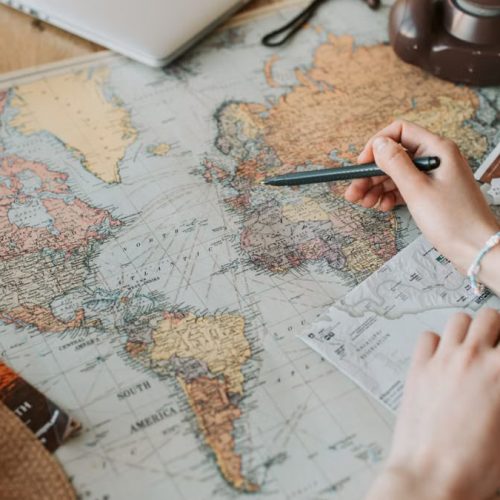 Hands marking destinations on a world map surrounded by travel essentials like passport and camera.
