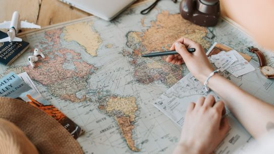 Hands marking destinations on a world map surrounded by travel essentials like passport and camera.